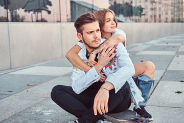 Trendy dressed young couple - pretty girl embrace her boyfriend while they sitting together on a skateboard near skyscraper.