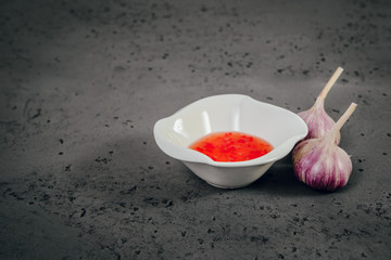 A view of a white bowl with red sauce and garlic next to it. Red sauce and garlic on a stone, dark table. The concept of preparing dishes, illustrative photo for food photography.