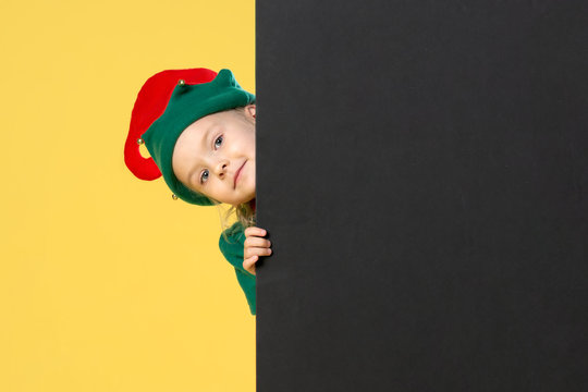 Little girl in a Christmas elf costume on a yellow background. A child looks out from behind a black sheet. Festive photo with place for text.