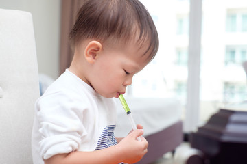 Cute little Asian 30 months / 2 years old toddler baby boy child who having flu feeding himself with liquid medicine with a syringe, Fever cold and flu concepts. Selective focus at medicine & syringe