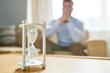Closeup of hourglass on table with blurred shape of man waiting in background, copy space