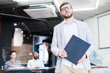 Content confident young bearded speaker of business training holding clipboard with thesis and looking at camera in conference room