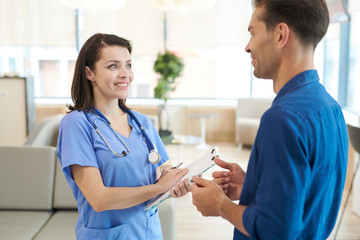 Waist up portrait of smiling young nurse talking to mature patient standing in hall of modern...