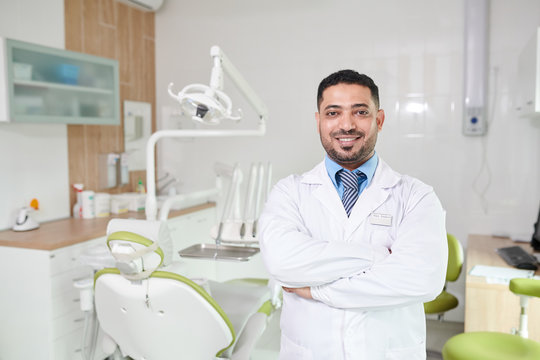 Waist up portrait of Middle-Eastern dentist posing in office smiling at camera while standing with arms crossed by dental chair, copy space