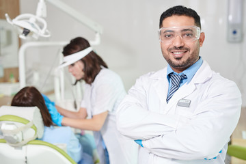 Obraz na płótnie Canvas Waist up portrait of confident Middle-Eastern doctor wearing lab coat posing in dentists office with patient in background, copy space