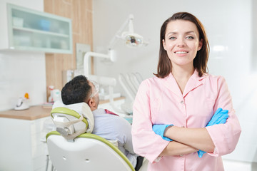 Waist up portrait of pretty female dentist looking at camera and smiling while posing standing with arms crossed in office, patient in background, copy space