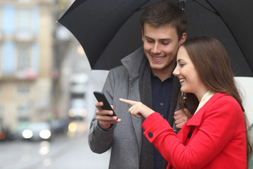 Happy couple checking phone in winter under rain
