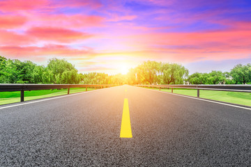 Empty asphalt road and green forest with colorful clouds at sunset