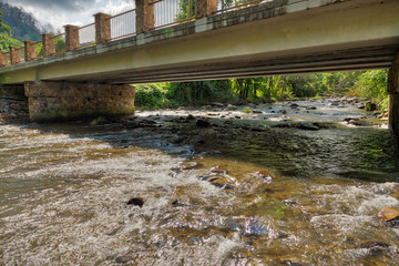 Rapidly flowing river going under a bridge with the sun reflecting brightly off the water and rocks
