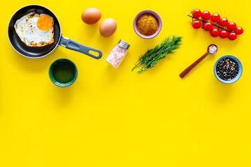 Recipe of fried eggs with vegetables. Ready eggs in a frying pan near cherry tomatoes, greenery, spices, raw eggs on yellow background top view copy space