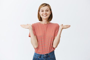 So waht I do not care. Portrait of careless happy and carefree attractive young female with blond haircut, shrugging with spread palms near shoulders, smiling joyfully, being unaware and uninterested