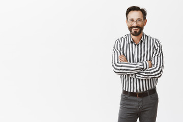 Fototapeta premium Attractive successful businessman in formal striped shirt and pants, holding hands crossed on chest and smiling broadly while standing on social evening, talking with business partners casually