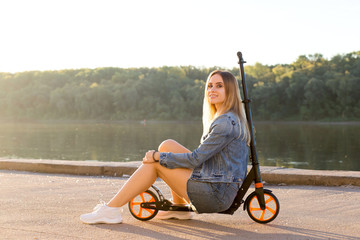 Beautiful sexy blonde woman in denim clothes sits on her scooter and smiles at the camera