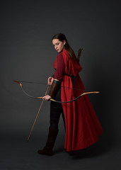 full length portrait of brunette girl wearing red medieval costume and cloak, holding a bow and...