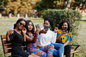 Group of four african american girls sitting on bench outdoor with mobile phones at hands and making selfie.