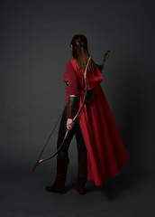 full length portrait of brunette girl wearing red medieval costume and cloak, holding a bow and arrow. standing pose on grey studio background.