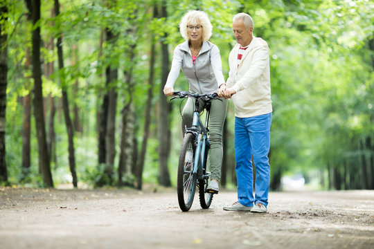 Mature woman sitting on bicycle and moving while her husband walking near by and supporting her