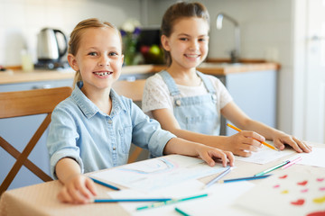 Obraz premium Two cheerful little girls in casualwear sitting by table in the kitchen while going to draw