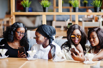 Four african american girls sitting on table at cafe with ice cream dessert and looking photos at mobile phones.