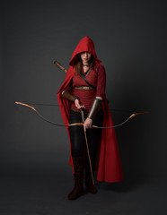 full length portrait of brunette girl wearing red medieval costume and cloak, holding a bow and...