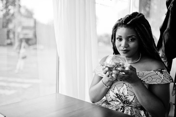 Cute small height african american girl with dreadlocks, wear at coloured yellow dress, sitting at cafe with flower at glass. Black and white.