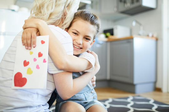 Smiling little girl with handmade greeting card embracing her mom in the kitchen