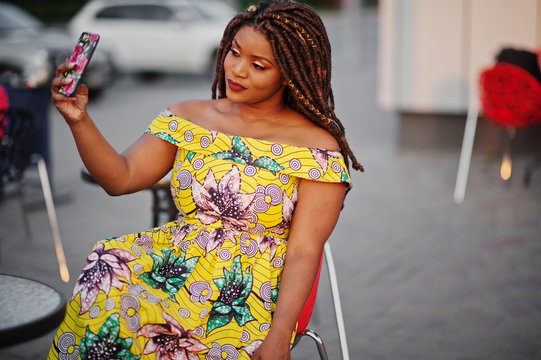 Cute small height african american girl with dreadlocks, wear at coloured yellow dress, sitting at outdoor cafe on red chair and making selfie.