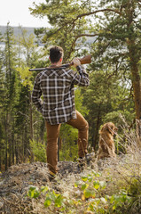 A hunter man staying in a forest and holding a gun on his shoulder and a dog