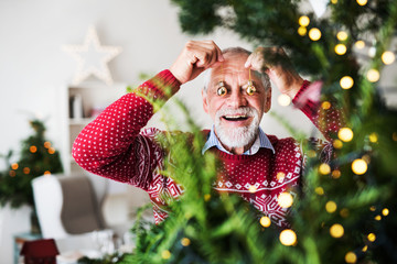 A senior man standing by Christmas tree, putting balls in front of eyes.