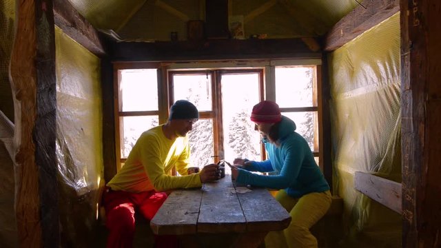 Happy couple of travelers is sitting at a wooden table against a window in frosty patterns, drinking coffee and using tablet pc. Awesome morning in old mountain shelter during a winter adventure.