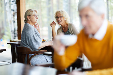 Content attractive mature ladies in casual clothing sitting at table and gossiping about handsome...