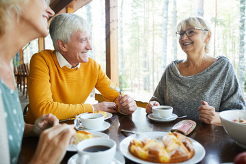 Cheerful excited senior friends in casual clothing sitting at table and drinking tea while sharing...