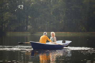 Smiling beautiful curly-haired senior lady in cardigan sitting on boat and talking to boyfriend while he rowing oars