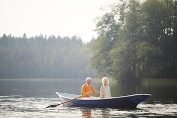 Excited handsome senior boyfriend in yellow sweater sitting on boat with beloved woman and rowing...
