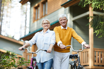 Cheerful active senior couples in casual clothing standing against country house and holding...