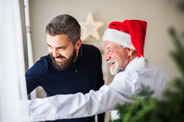 Obraz premium A senior father with a Santa hat and adult son standing by the window at home.
