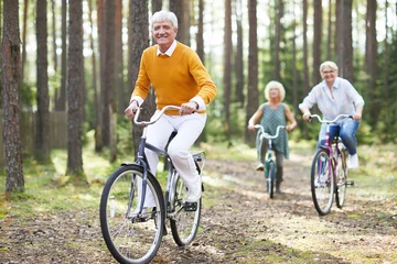 Fotobehang Cheerful excited senior friends in casual clothing enjoying active life cycling on bikes together in forest © pressmaster