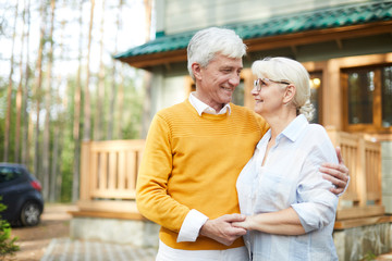 Positive romantic senior couple embracing and holding hands while looking into eyes, they standing against cottage