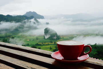Hot coffee latte cappuccino in red cup on wooden terrace with beautiful scenic view nature background of misty morning mountain in Phayao, Thailand.