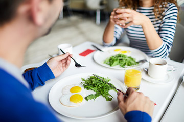 Close-up of unrecognizable man holding silverware and eating fried eggs with greens at breakfast with friend in cafe