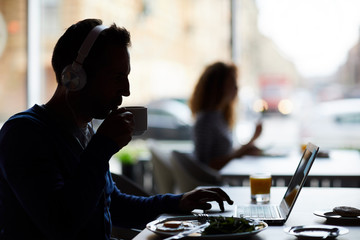 Silhouette of male hipster entrepreneur in headphones sitting at table and using laptop while drinking coffee and working in cafe