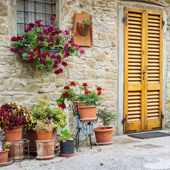 Fototapeta na wymiar Beautiful flowers in front of stone wall in a small village of medieval origin. Volpaia, Tuscany, Italy. Square shape