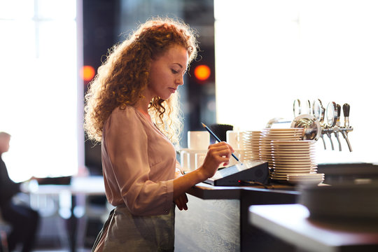 Serious pensive attractive curly-haired waitress standing at bar counter and using pencil while adding order in restaurant POS