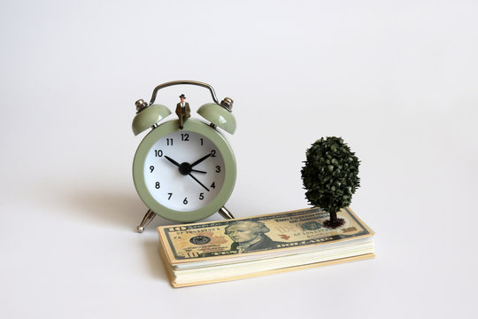 A miniature man sitting on the an alarm clock and dollar.