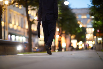 Businessman with briefcase walking down street in the evening along green trees