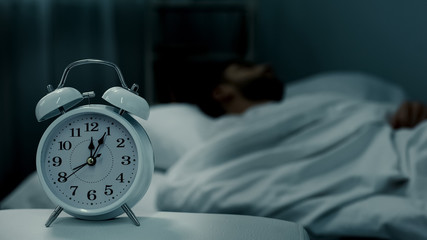 Man sleeping in bed, alarm clock showing midnight, rest hours, healthy sleep