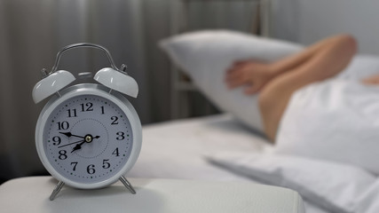 Sleepy female annoyed by ringing alarm clock closing ears with pillow, jet lag