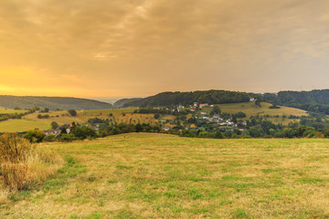 Eifel landscape at sunrise with view on village Udscheid against background with morning fog and hills with forests and sky with light clouds