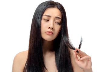 portrait of upset asian woman looking at split ends isolated on white, damaged hair concept