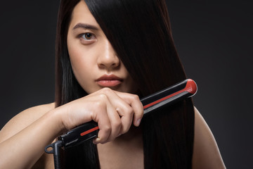 portrait of young asian woman straightening hair with hair straightener isolated on black